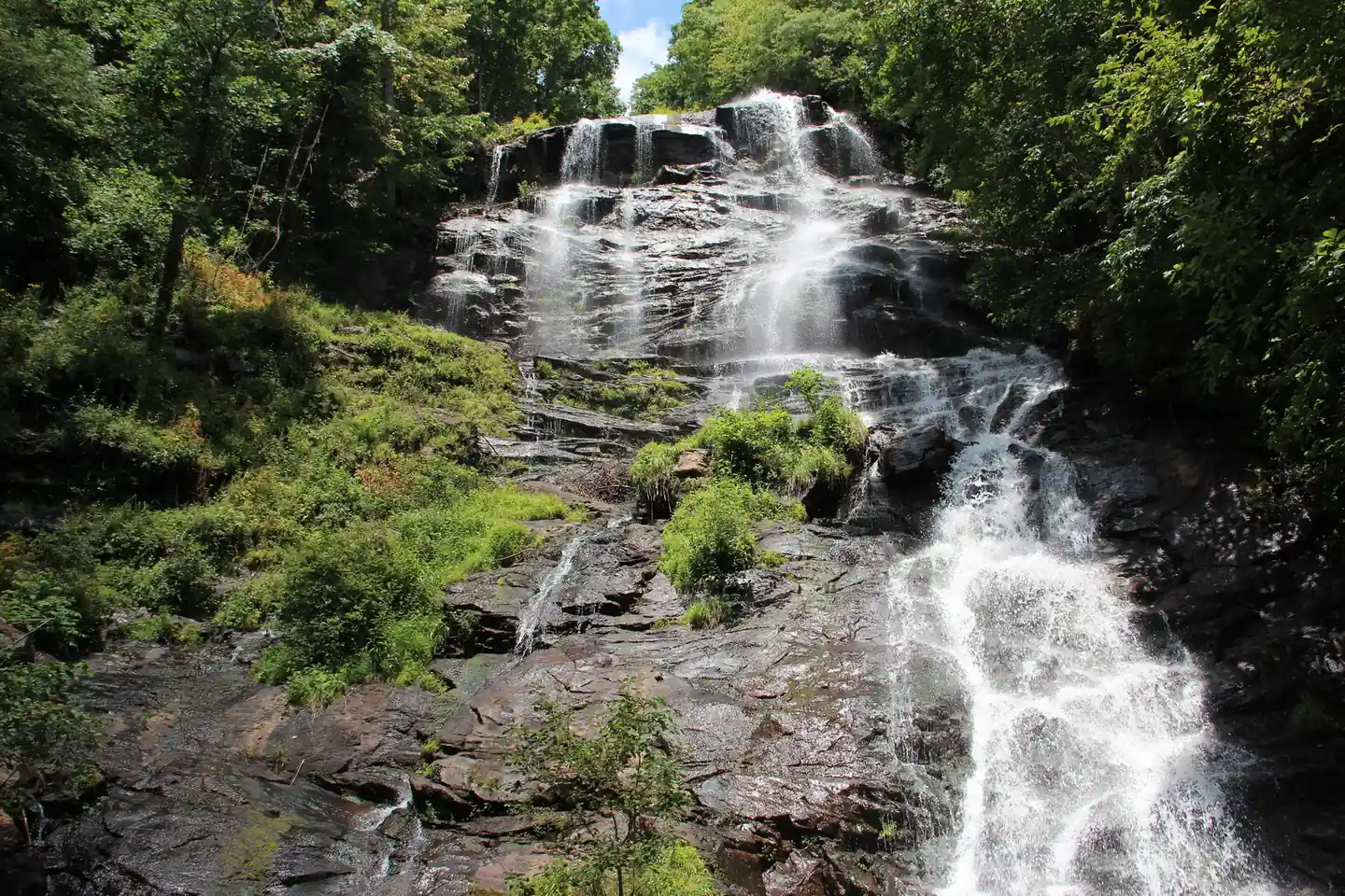 Looking up at Amicalola Falls in summer: white water fanning down a tall dark rock face, with a lower torrent pouring down the right side, green shrubs on the rocks, dense forest on both sides and a sliver of blue sky at top.