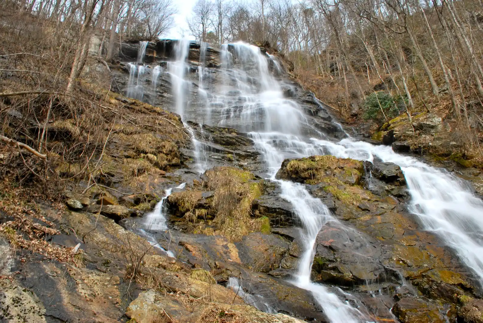 Long-exposure view of a big multi-channel cascade pouring down sloping rock among bare late-winter trees, with mossy and golden lichen-covered boulders in the foreground. Bright white water, hazy white sky at top.