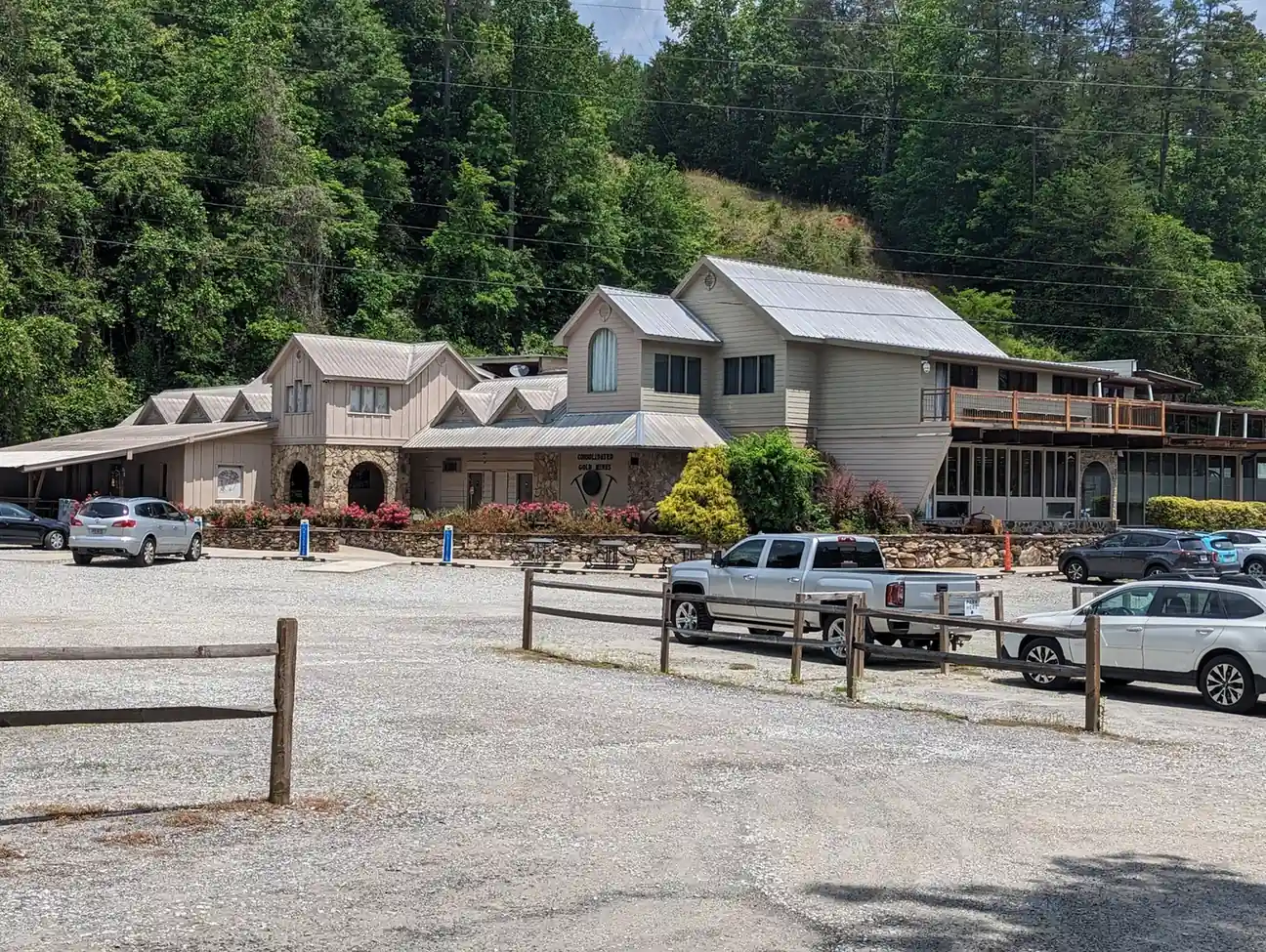 Exterior of the Consolidated Gold Mine visitor building: a long tan two-story structure with metal roofs, stone arches and a deck, set against a steep forested hillside; gravel parking lot with a silver pickup, SUVs and split-rail fence in front; power lines across the top. No people visible.