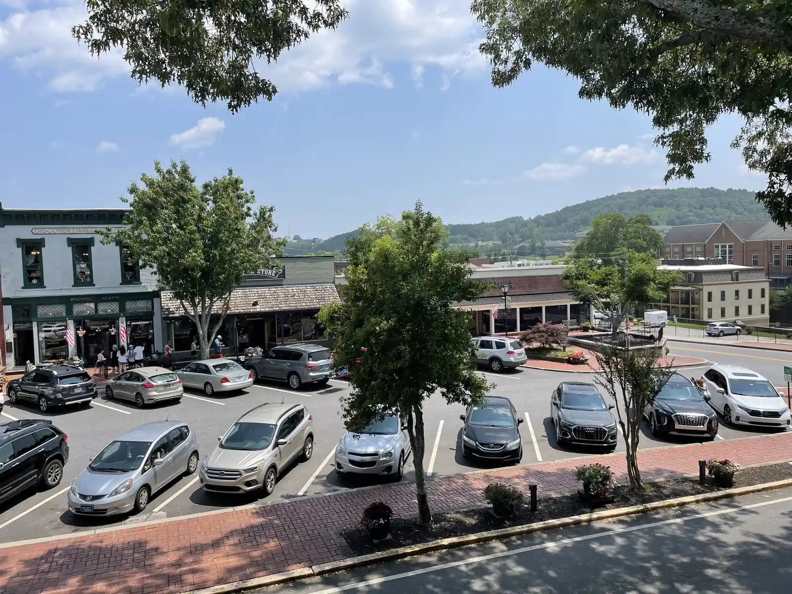 Summer view looking down from the Gold Museum's upper level across the square's parking lot: a row of 19th-century two-story commercial buildings (green-trimmed facade at left, low shingled shopfronts center), about 15 parked cars, young street trees, brick sidewalk in the foreground, and a wooded green ridge rising behind the town under blue sky with cumulus. A few tiny pedestrians by the shops; no legible signs.