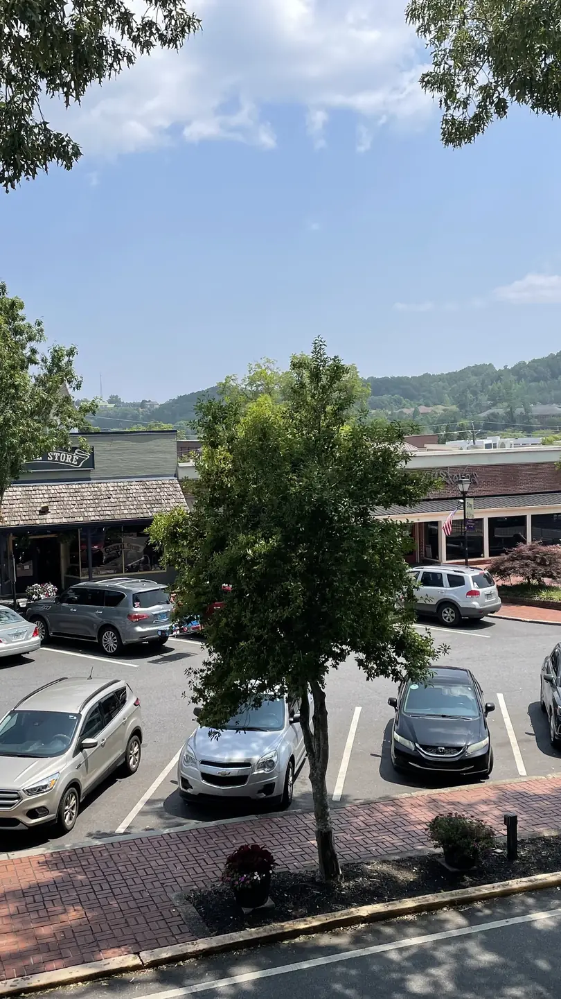Summer view looking down from the Gold Museum's upper level across the square's parking lot: a row of 19th-century two-story commercial buildings (green-trimmed facade at left, low shingled shopfronts center), about 15 parked cars, young street trees, brick sidewalk in the foreground, and a wooded green ridge rising behind the town under blue sky with cumulus. A few tiny pedestrians by the shops; no legible signs.