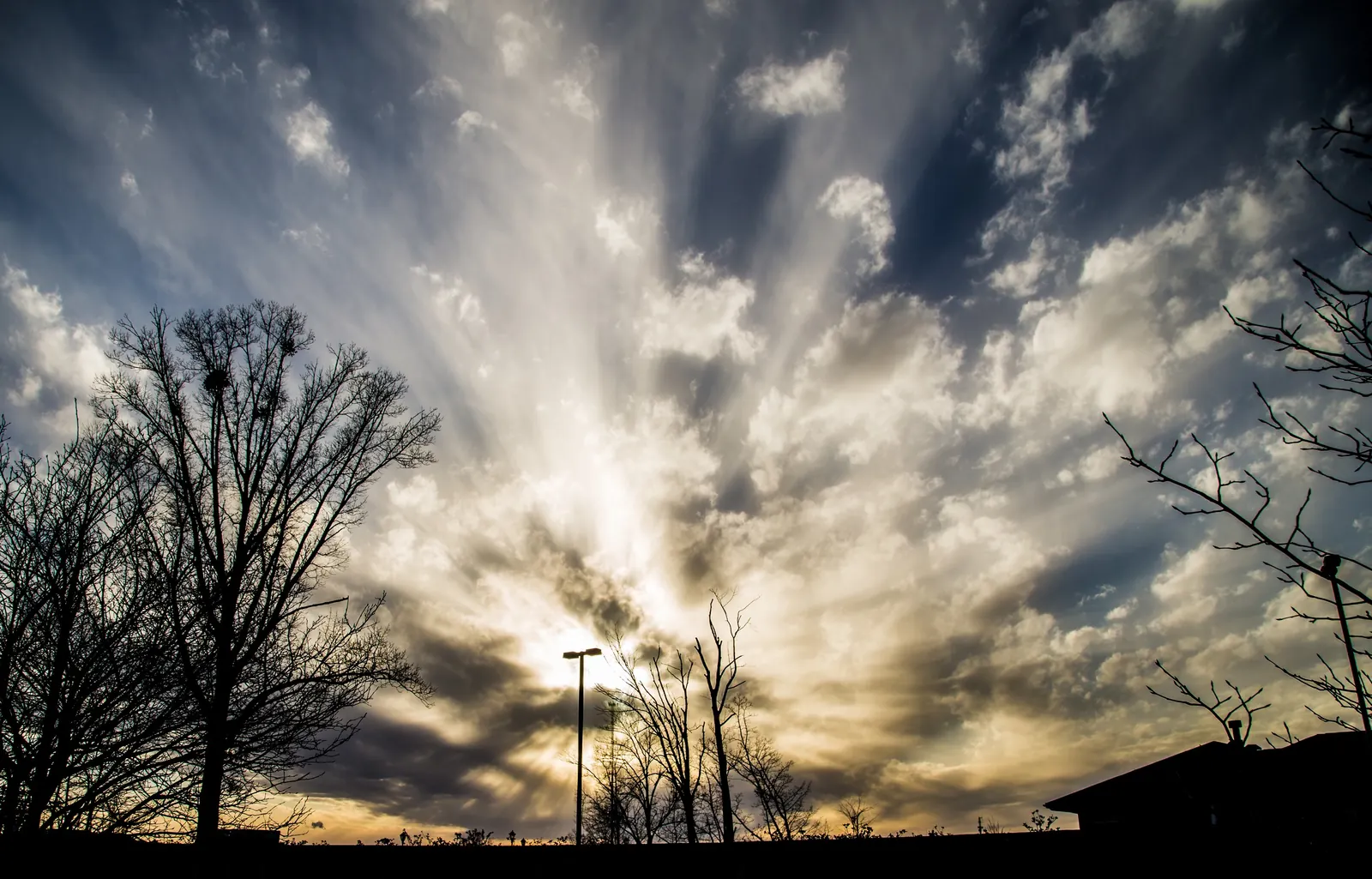 Dramatic winter sunset: crepuscular rays fanning through broken clouds over silhouetted bare trees, a single street lamp and the dark roofline of a building at bottom right; ground is black silhouette.