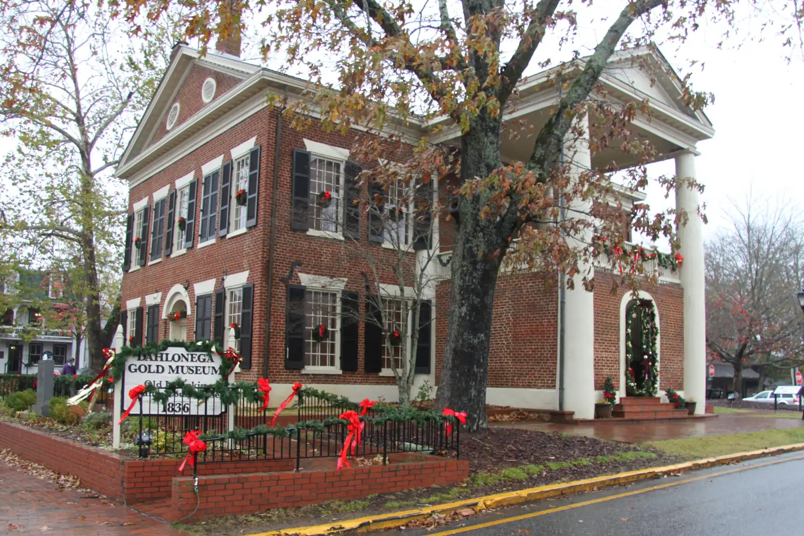 Three-quarter view of the Gold Museum's brick side and white columned portico on a wet grey day, with a large oak in front, holiday garland and red ribbons on the iron fence and on the 'Dahlonega Gold Museum - Old Courthouse - 1836' sign at left, rain-dark street curving along the foreground. No people.