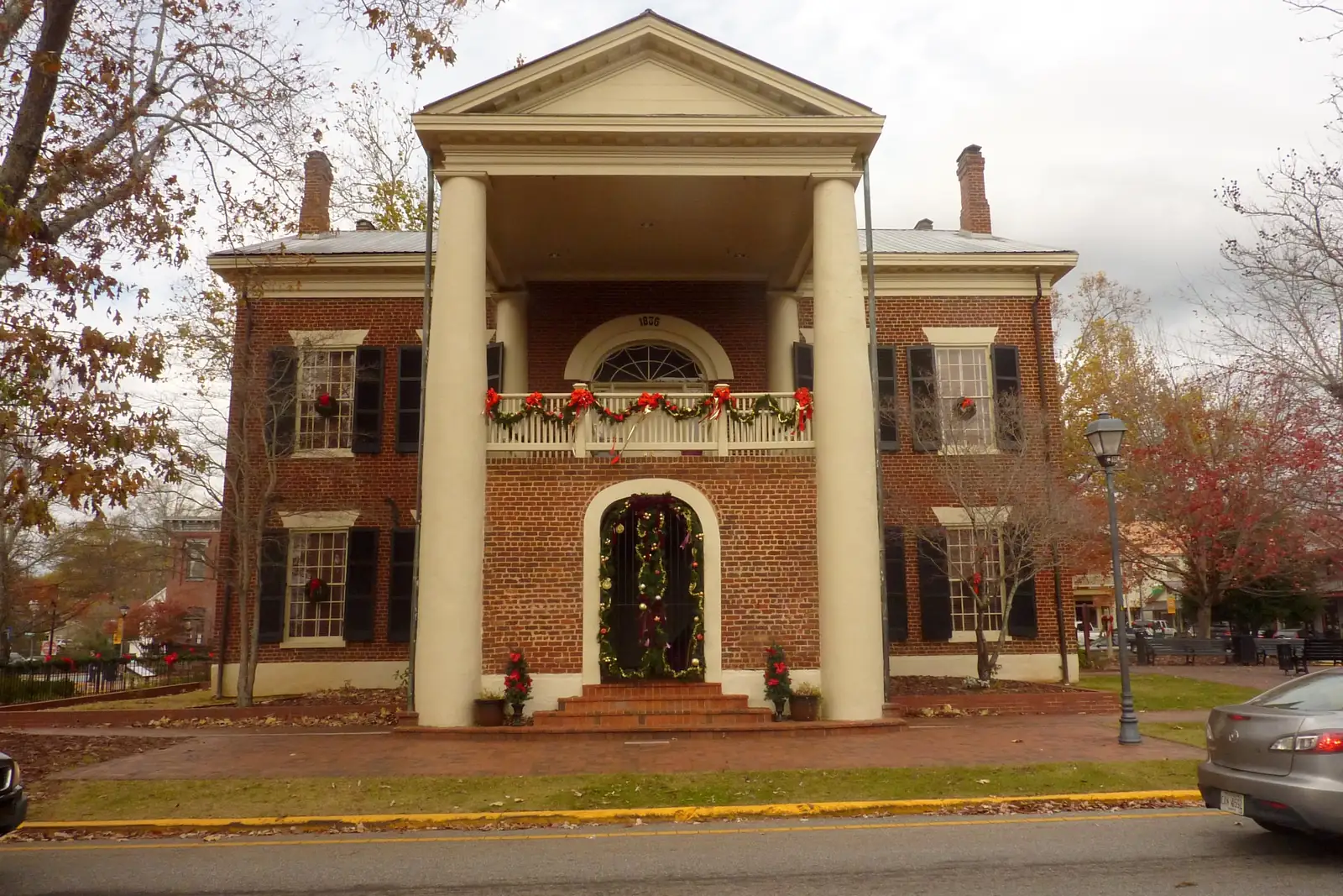 Front of the red-brick, white-columned Gold Museum decorated for Christmas: evergreen garland with red bows on the balcony rail, a wreath-framed arched doorway, red flowers by the steps. Autumn trees with russet and red leaves on both sides, overcast sky, street and yellow curb in the foreground, the rear of a silver car cut off at right edge.