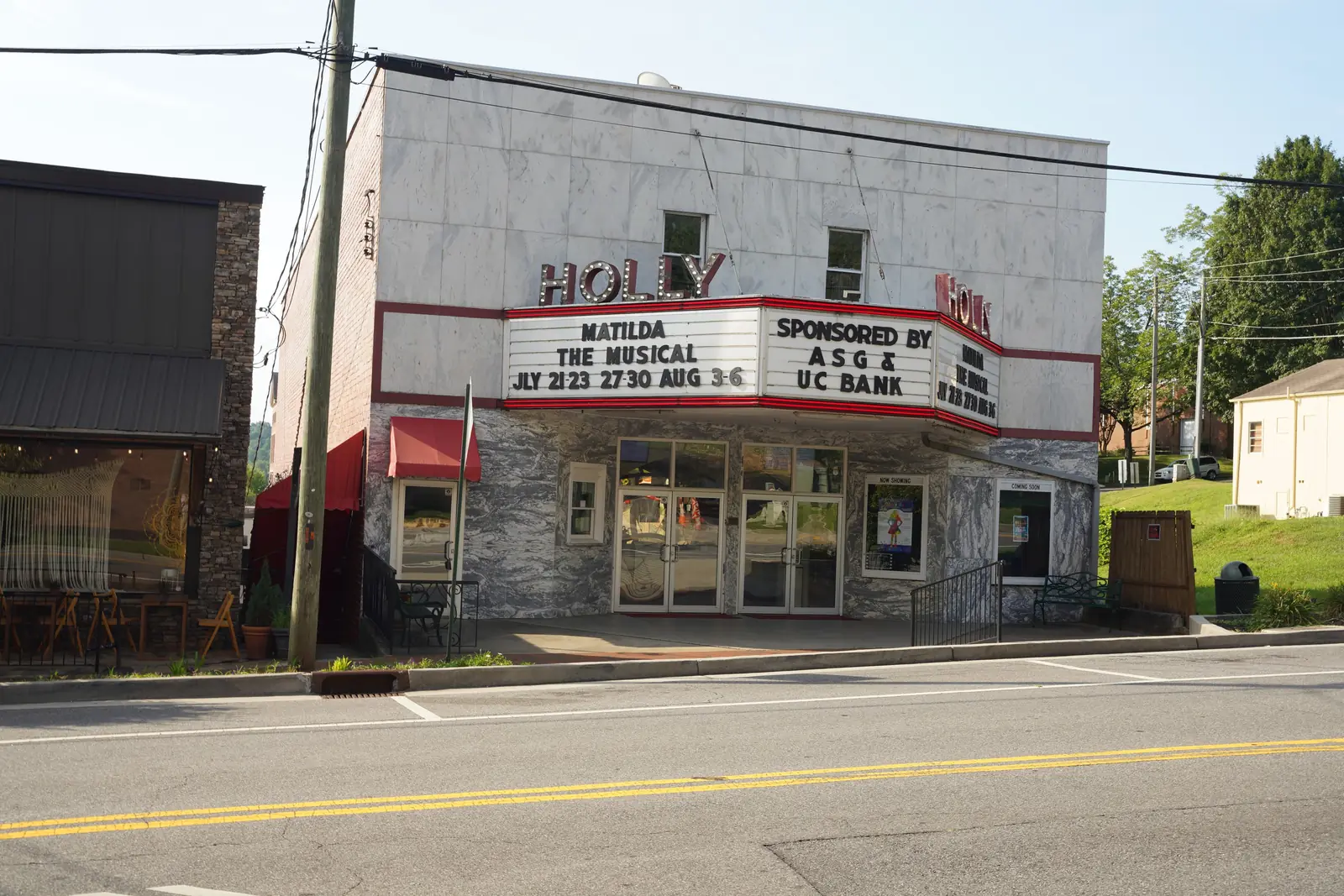 Front of the Holly Theater: white marble-clad two-story facade with red-outlined 'HOLLY' letters and a marquee reading 'MATILDA THE MUSICAL ... SPONSORED BY ASG & UC BANK', glass entry doors, a utility pole and wires at left, part of a neighboring dark storefront at left, empty street in front, summer trees behind. No people.