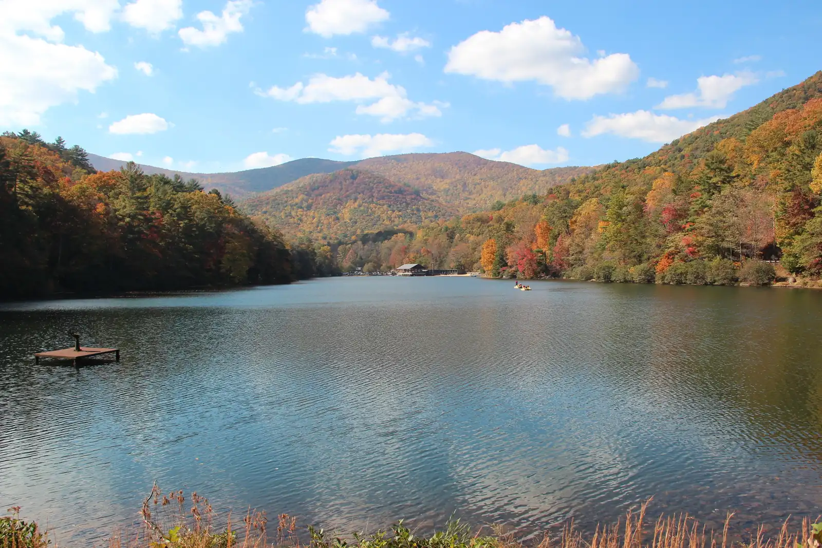 Classic Vogel view across Lake Trahlyta: rippled blue water reflecting the sky, a small floating dock at left, the lakeside pavilion in the distance, one small boat with tiny figures mid-lake, and fall-colored mountains (orange, gold, green) rising behind under a blue sky with white cumulus. Dry grasses along the bottom edge.