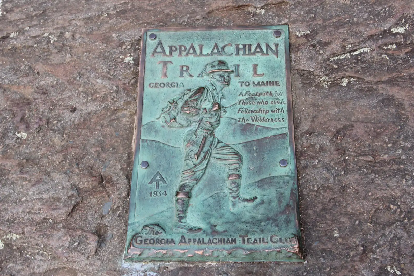 Close-up of the bronze/verdigris Appalachian Trail plaque set in rock: relief of a hiker with pack, text 'Appalachian Trail - Georgia to Maine - A Footpath for Those who seek Fellowship with the Wilderness - 1934 - The Georgia Appalachian Trail Club'.
