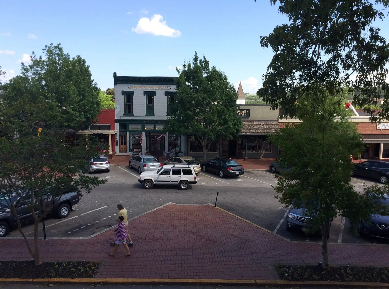 Looking down from the Gold Museum balcony at the square in summer: a white two-story 1800s storefront with green trim, neighboring shops, a parking lot with about eight cars including a white Jeep, brick crosswalk, and two pedestrians (small, mid-frame, walking away/sideways). Trees frame both sides, blue sky with one white cloud.